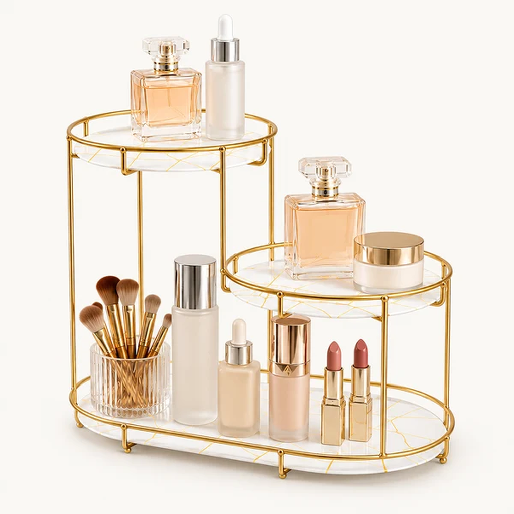 A two-tier gold metal and white marble makeup organizer styled with various cosmetic products, including bottles, lipsticks, brushes, and perfume, on a clean white background.