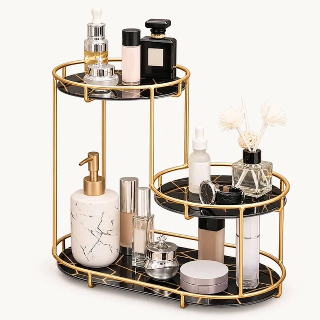 A gold-framed two-tier bathroom vanity organizer with black marble-look trays, styled with cosmetic bottles, jars, and a reed diffuser on a light off-white background.
